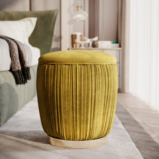 A Ruched Pouffe With Gold Plated Base in mustard yellow is positioned next to a green-upholstered bed. This elegant piece enhances the bright, modern bedroom that features neutral-toned walls and sheer white curtains, allowing natural light to filter in.