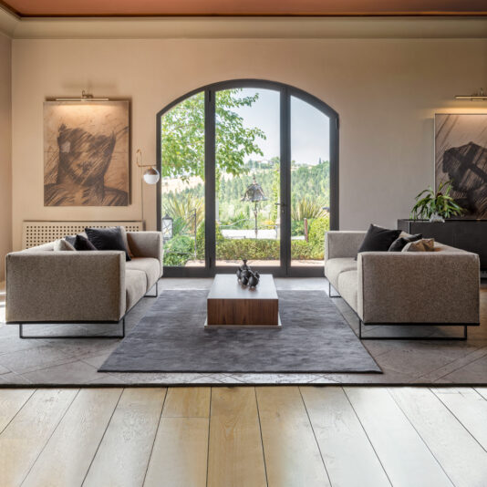 Walnut Veneer Box Style Storage Coffee Table A modern living room with two gray sofas facing each other, featuring a Walnut Veneer Box Style Storage Coffee Table in between them. Large arched glass doors open to a lush, green outdoor space. The walls are adorned with two framed abstract artworks, and a floor lamp stands in the corner.