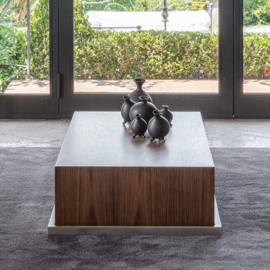 A Walnut Veneer Box Style Storage Coffee Table with a minimalist, modern design sits on a dark carpet in front of large glass doors. The table is adorned with several black ceramic vases of varying shapes and sizes, arranged together in a cluster. A green hedge is visible outside the doors.