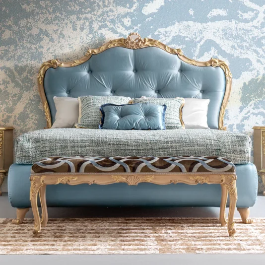 The Button Upholstered Rococo Style Bed features a tufted light blue headboard with ornate golden trim and comes with decorative pillows and a textured blanket. A matching upholstered bench with gold accents is at the foot, set against a blue textured wall backdrop.