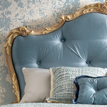Button Upholstered Rococo Style Bed The Button Upholstered Rococo Style Bed boasts a tufted blue headboard with ornate gold detailing, adorned with white, light gray, and textured blue pillows against blue and white patterned wallpaper.