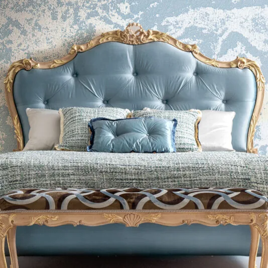 Button Upholstered Rococo Style Bed A luxurious bedroom features a large Rococo-style headboard with button upholstery. The bed is adorned with textured blue and white pillows, complemented by a patterned blanket, set against a blue and white textured wall. Product: Button Upholstered Rococo Style Bed.