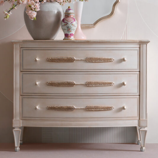 The Classic Feather Design Chest Of Drawers boasts a vintage style with three drawers, each adorned with gold leaf-shaped handles. On top, decorative items include a vase with pink flowers, a pink and white ceramic jar, and a small pink object. A partial mirror is visible in the background.