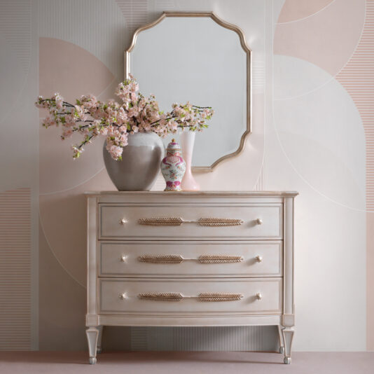 The Classic Feather Design Chest Of Drawers, with its three compartments and elegant gold accents, stands against a chic light-colored geometric wallpaper. Two large vases filled with pink flowers and a smaller decorative vase adorn the top of the dresser, while an ornate mirror hangs above it.