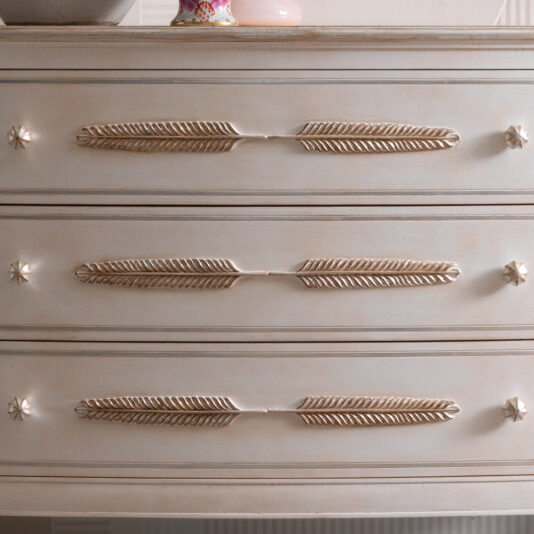 A close-up of the vintage Classic Feather Design Chest Of Drawers showcasing its three drawers, each adorned with decorative gold feather-shaped handles and star-shaped knobs. The dresser boasts a charming light, distressed finish.
