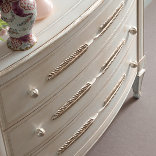 A close-up view of the Classic Feather Design Chest Of Drawers in vintage white showcases its ornate carvings on the drawers. This elegant dresser features three drawers with decorative handles and intricate detailing. A portion of a porcelain vase with floral patterns is visible on the top left corner.
