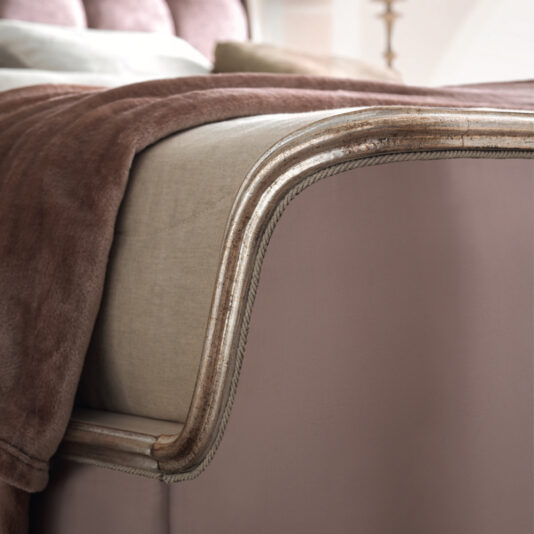 Close-up of the Feather Design Button Upholstered Sleigh Bed featuring a curved, gold-trimmed headboard and footboard. The bed is adorned with beige and dusty pink bedding, including a soft, plush blanket draped over the side. The overall aesthetic is elegant and sophisticated.