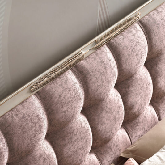 A close-up of the Feather Design Button Upholstered Sleigh Bed showcases an elegant tufted headboard in soft pink fabric with metallic trim and decorative accents at the top. The headboard, adorned with feather design buttons, exhibits a luxurious, textured appearance and is part of a neatly made bed with matching bedding.