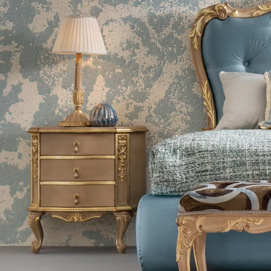 In a luxurious bedroom corner, an ornate Classic Style Bedside Cabinet with gold accents is adorned by a decorative lamp and ornament. Beside it, a bed features an upholstered blue headboard and textured bedding. A patterned stool sits in front, complemented by elegant wallpaper.