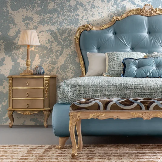 An elegant bedroom showcases a luxurious blue and gold bed with an ornate headboard. Beside the bed rests the Classic Style Ornate Bedside Cabinet with a matching lamp. The wall features a textured pattern, and a cushioned bench with intricate designs is at the foot of the bed.
