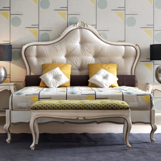 A stylish bedroom features the Classic Style Ornate Button Upholstered Bed, with its plush, tufted headboard that embodies a classic style. The bed is adorned with yellow and white pillows and a patterned blanket. Two matching bedside tables with lamps add symmetry, while the wallpaper showcases a geometric pattern in soft colors.