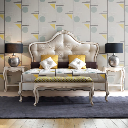 A stylish bedroom showcases the Classic Style Ornate Button Upholstered Bed with its elegant beige headboard. The bed is decorated with pillows and bedding in geometric patterns, featuring hues of yellow, white, and gray. Flanking the bed are two exquisite bedside tables, each topped with a metallic lamp. A bench at the foot of the bed enhances the timeless ambiance. All set against a backdrop of modern geometric wallpaper, this bedroom epitomizes classic sophistication.