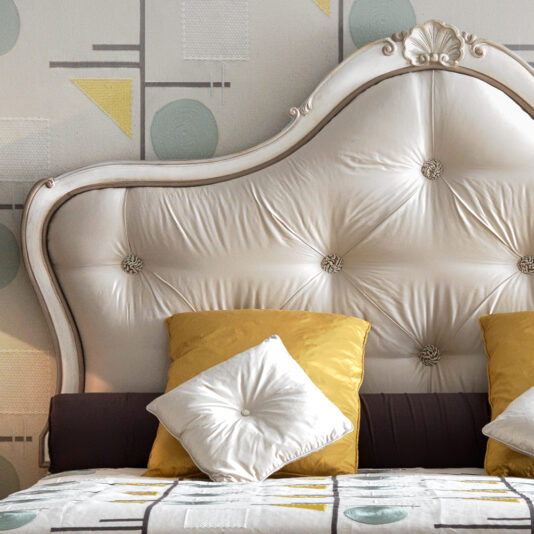 Image of a luxurious bedroom with a Classic Style Ornate Button Upholstered Bed featuring a tufted headboard in white. The bed is adorned with mustard yellow pillows, a white cushion in front, and a patterned blanket with circular and rectangular motifs. The wall showcases geometric-patterned wallpaper in light pastel colors.