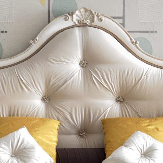 The Classic Style Ornate Button Upholstered Bed in a cream color provides an elegant backdrop with its tufted button details. On the bed, two mustard yellow pillows and two white pillows are stylishly arranged. The background wallpaper showcases a geometric pattern in subtle tones.
