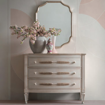 Curved Italian Wall Mirror A stylish dresser with three drawers adorned with feather-shaped handles stands against a pastel geometric-patterned wall. On top of the dresser are a gray vase with pink blossoms, a decorative jar, and a pink vase. Above, the Curved Italian Wall Mirror in a gold frame hangs gracefully.