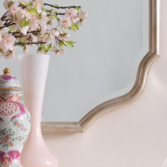 A decorative scene featuring a pink and white floral vase with intricate designs, holding blooming branches with pink flowers. The vase is placed beside the Curved Italian Wall Mirror, which has a large frame accented with gold details. The background is a soft beige wall.