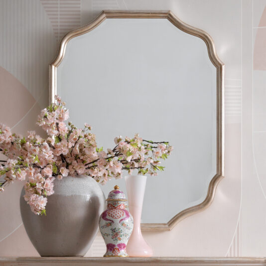 A decorative Curved Italian Wall Mirror with an ornate, scalloped gold frame hangs above the mantel. On the mantel, there is a large beige vase filled with pink cherry blossoms, a decorative porcelain jar with a floral pattern, and a simple white and pink vase.