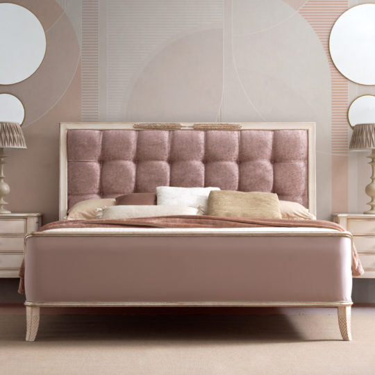 A stylish bedroom features the Feather Design Button Upholstered Sleigh Bed with a cushioned, tufted headboard in dusty rose. The bed is adorned with multiple pillows in varying shades of tan and white. Two matching nightstands with table lamps flank the bed, and round mirrors hang above them.