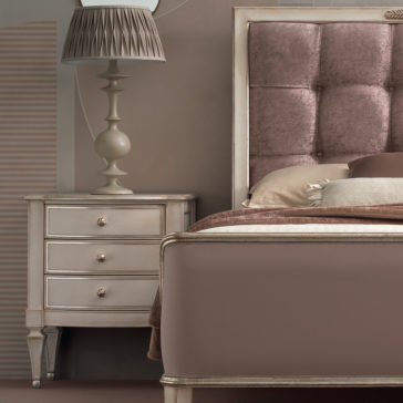 High End 3 Drawer Bedside Cabinet A cozy bedroom scene featuring the edge of a taupe-colored bed with a tufted velvet headboard. Beside the bed, there is a High End 3 Drawer Bedside Cabinet adorned with an ornate lamp that has a pleated lampshade. The overall color scheme is soft, neutral tones.