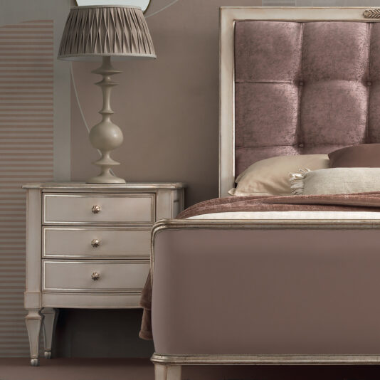 A cozy bedroom scene featuring the edge of a taupe-colored bed with a tufted velvet headboard. Beside the bed, there is a High End 3 Drawer Bedside Cabinet adorned with an ornate lamp that has a pleated lampshade. The overall color scheme is soft, neutral tones.