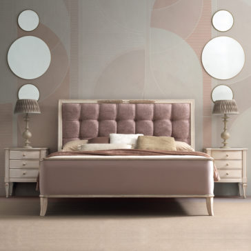 High End 3 Drawer Bedside Cabinet A modern bedroom features a bed with a padded, tufted headboard in soft pink fabric, flanked by High End 3 Drawer Bedside Cabinets. Each cabinet holds a lamp with a pleated shade. The wall has an abstract design adorned with four round mirrors.