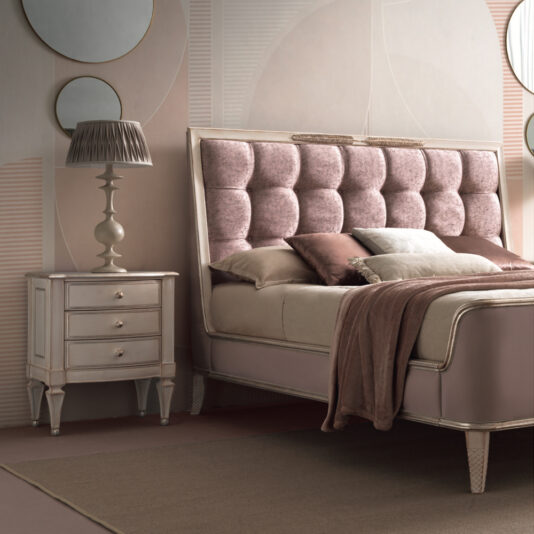 High End 3 Drawer Bedside Cabinet A luxurious bedroom features a bed with a tufted pink and beige headboard, matching linens, and a plush throw blanket. To the left of the bed stands the High End 3 Drawer Bedside Cabinet, topped with a decorative lamp. The walls showcase geometric patterns in soft hues.