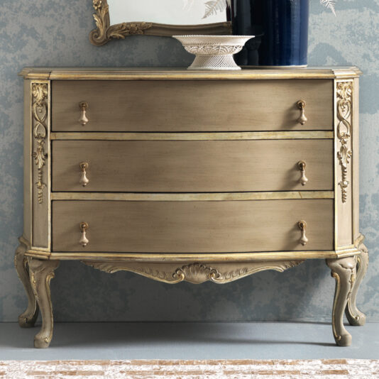 The Ornate Classic Style Chest Of Drawers, an antique piece in beige, features three compartments adorned with brass handles and intricate floral carvings on the sides. It stands proudly on ornate, curved legs against a textured blue wall, topped with a decorative bowl and a blue vase.