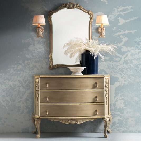 The Ornate Classic Style Chest Of Drawers, featuring three compartments, stands against a textured blue wall. Above it hangs an elegant mirror with a decorative frame, flanked by two wall sconces with lamps. A dark vase filled with white feathery branches and a white bowl adorn the dresser top.