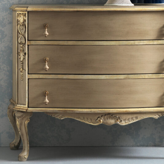 The Ornate Classic Style Chest Of Drawers boasts an antique finish, intricate floral carvings, brass handles, and elegantly curved legs. Its distressed wood enhances its classical charm against a muted blue wall backdrop.