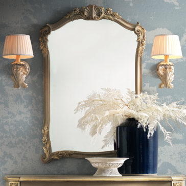 Elegant vintage-style bathroom mirror featuring an Ornate Classic Style Wall Mirror with a gold frame, flanked by two matching wall sconces with pleated shades. Beneath the mirror, a dark blue vase holds white botanical arrangements, standing next to a decorative dish, all on a narrow console table.