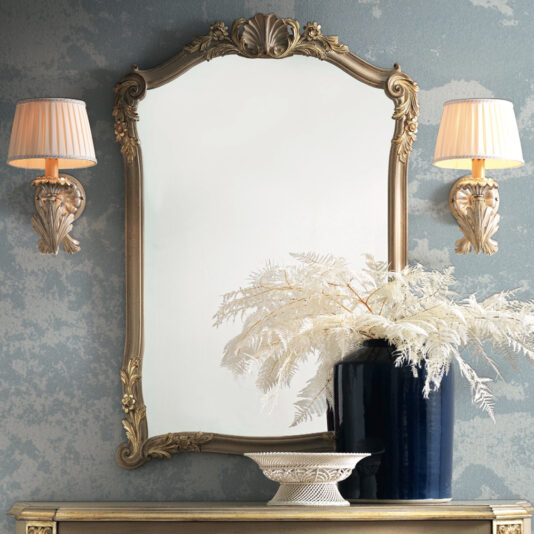 Elegant vintage-style bathroom mirror featuring an Ornate Classic Style Wall Mirror with a gold frame, flanked by two matching wall sconces with pleated shades. Beneath the mirror, a dark blue vase holds white botanical arrangements, standing next to a decorative dish, all on a narrow console table.
