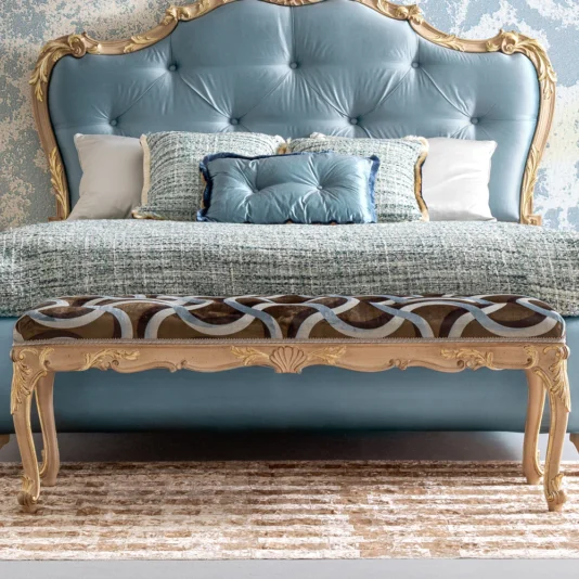 A luxurious bedroom features a tufted blue headboard with decorative pillows and a blue-and-white textured bedspread. An Ornate Rococo Style Bench with intricate gold details sits at the bed's foot on a plush, light-colored rug.