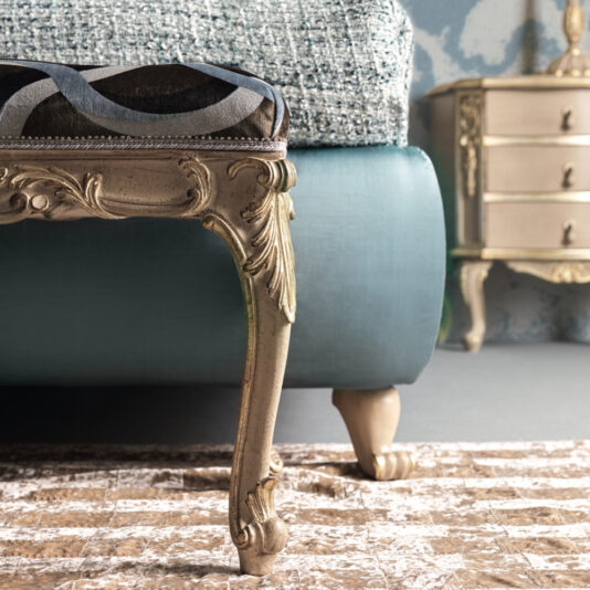 Ornate Rococo Style Bench A close-up of an intricately carved leg of the Ornate Rococo Style Bench, showcasing its vintage design and elaborate details. The bench is upholstered in teal fabric, and the foreground displays a patterned carpet. In the background, there is a vintage bedside table.