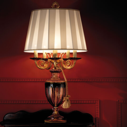 The Candelabra Style Amber And Blue Glass Table Lamp, with a striped white lampshade and an ornate gold base, sits on a dark wood table against a deep red wall. The lamp features multiple candle-like bulbs, intricate gold detailing, amber and blue glass accents, and a gold tassel.