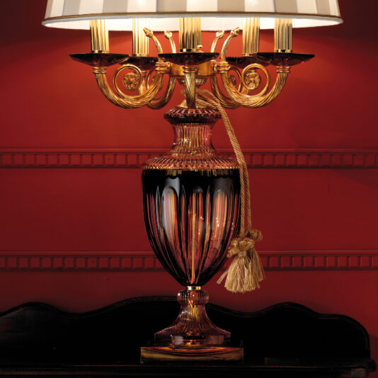 The Candelabra Style Amber And Blue Glass Table Lamp features an ornate, gold-colored base and a unique maroon and blue glass body. It boasts an intricate design with curved arms holding multiple candle-like light bulbs, a beige and white striped lampshade, and a tassel hanging from its neck.