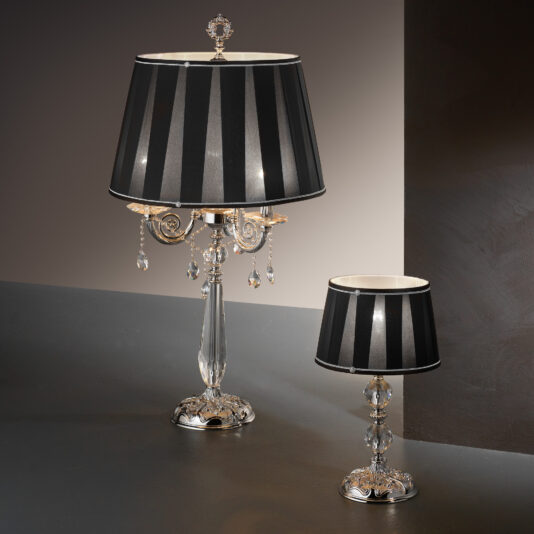 Two elegant "Candelabra Style Table Lamps With Swarovski Crystals" sit on a gray surface against a dark, gradient background. The larger lamp showcases a crystal base and ornate metallic detailing adorned with hanging Swarovski crystals, while the smaller lamp mirrors these exquisite features. Both lamps are topped with chic black and white striped lampshades.