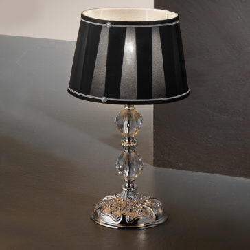 Candelabra Style Table Lamp With Swarovski Crystals The Candelabra Style Table Lamp With Swarovski Crystals features a metallic ornate base adorned with clear, faceted Swarovski crystal embellishments. It includes a black lampshade with vertical white stripes and subtle detailing at the connection point to the base. The lamp is displayed on a gray surface against a dark background.