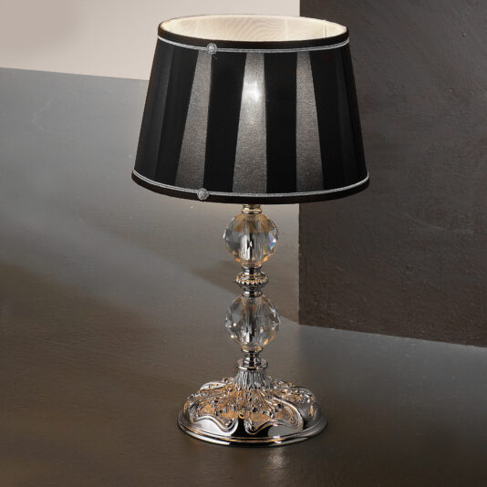 The Candelabra Style Table Lamp With Swarovski Crystals features a metallic ornate base adorned with clear, faceted Swarovski crystal embellishments. It includes a black lampshade with vertical white stripes and subtle detailing at the connection point to the base. The lamp is displayed on a gray surface against a dark background.