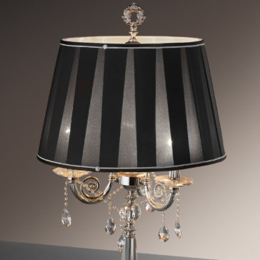 Candelabra Style Table Lamp With Swarovski Crystals A Candelabra Style Table Lamp With Swarovski Crystals, made of silver metal and adorned with a black and silver striped lampshade, showcases ornate detailing and Swarovski crystal pendants hanging below the bulb area. The lamp is turned off, set against a background gradient that shifts from dark to light brown.