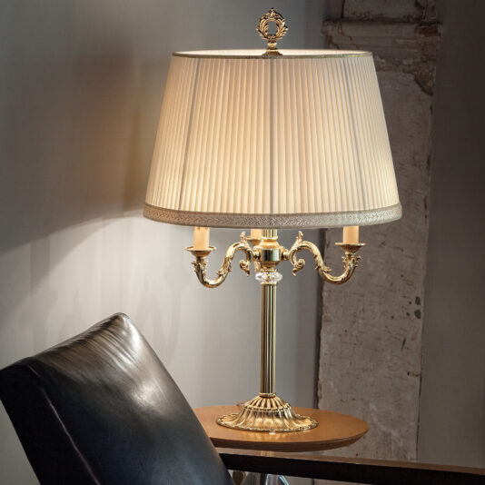 A Candelabra Style Table Lamp With Swarovski Detail, featuring an ornate gold base and topped with a pleated cream-colored shade, sits on a wooden side table next to a black leather chair. The lamp is turned on, casting a warm light, and is positioned against a plain wall with a portion of exposed column.