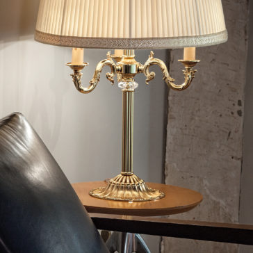 The Candelabra Style Table Lamp With Swarovski Detail, featuring a classic brass design and three arms, stands on a wooden side table next to a black leather chair. Its pleated beige lampshade softly illuminates the surrounding area, adding a touch of elegance to the room.