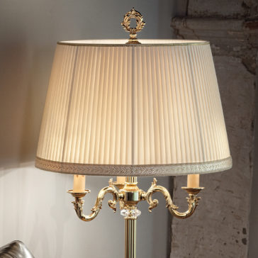 A Candelabra Style Table Lamp With Swarovski Detail and an ornate brass design stands against a rustic wall backdrop. It features a pleated white fabric shade and elegant double-arm holders with intricate detailing, casting a warm, inviting glow.