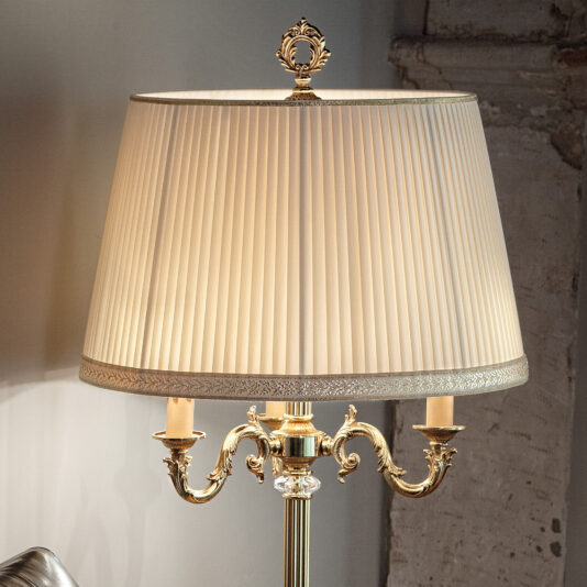 A Candelabra Style Table Lamp With Swarovski Detail and an ornate brass design stands against a rustic wall backdrop. It features a pleated white fabric shade and elegant double-arm holders with intricate detailing, casting a warm, inviting glow.