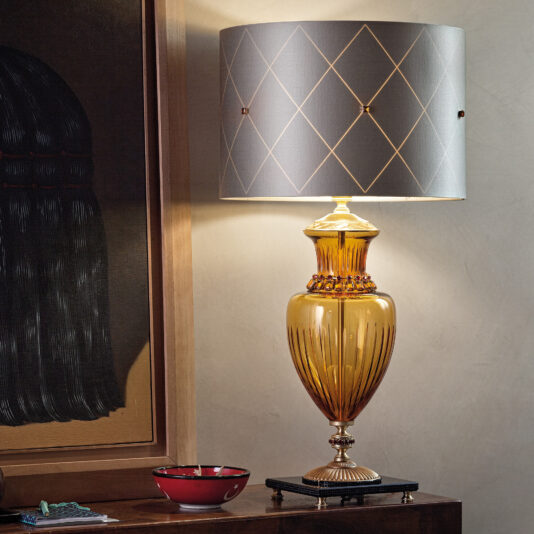 A stylish Classic Amber Glass Table Lamp With Amber Stones, adorned with a gray lampshade featuring a gold diamond pattern, is placed on a wooden surface. Nearby, a red and white ceramic bowl and a small patterned item are arranged. Part of dark wall art is visible in the background.