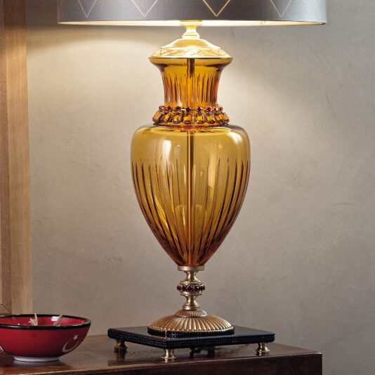 A Classic Amber Glass Table Lamp With Amber Stones, featuring intricate gold detailing and amber stones, stands elegantly on a polished dark wooden table. The lampshade is partially visible and has a black outer layer with a subtle design. Next to the lamp is a red bowl adorned with a crescent moon and star design.