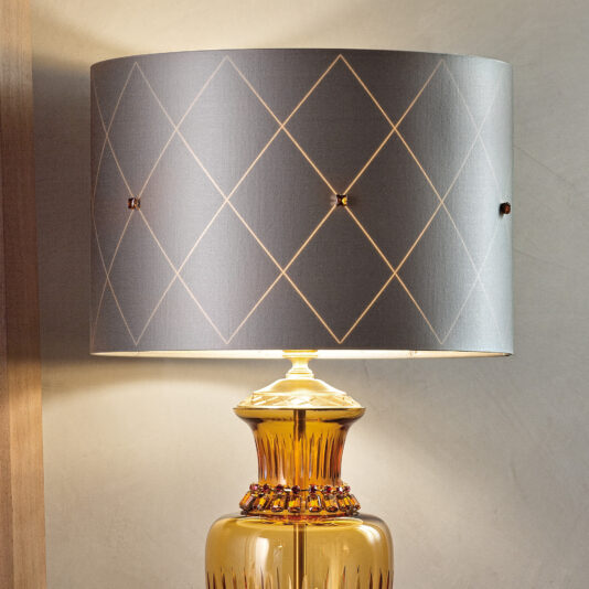 A close-up of the Classic Amber Glass Table Lamp With Amber Stones, showcasing its yellow, textured base embedded with amber stones. The lamp's cylindrical shade is adorned with gray fabric featuring a diamond pattern. The lamp is illuminated, casting a warm light against a plain wall in the background.