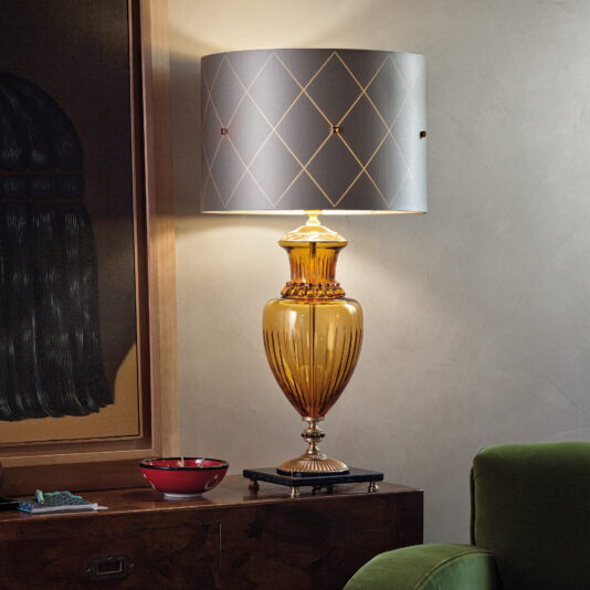 A beautifully decorated living space features the Classic Amber Glass Table Lamp With Amber Stones and a diamond-patterned grey lampshade. The lamp rests on a wooden cabinet beside a red bowl and a book, with a piece of artwork partially visible in the background.
