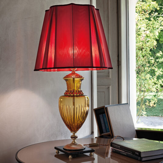A stylish Classic Amber Glass Table Lamp With Ruby Stones and a red fabric shade sits on a round wooden table next to a window. The table holds a book, a smartphone, a notepad, and a pen. Sunlight filters through the window, illuminating the amber glass base adorned with ruby stones.