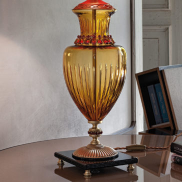 Classic Amber Glass Table Lamp With Ruby Stones A Classic Amber Glass Table Lamp With Ruby Stones, featuring an ornate amber glass body and a red-tinted top, sits on a decorative metal base atop a reflective surface. Behind it, there's a white wall and an open bookshelf filled with books. The lamp's elegant design enhances the scene's vintage aesthetic.
