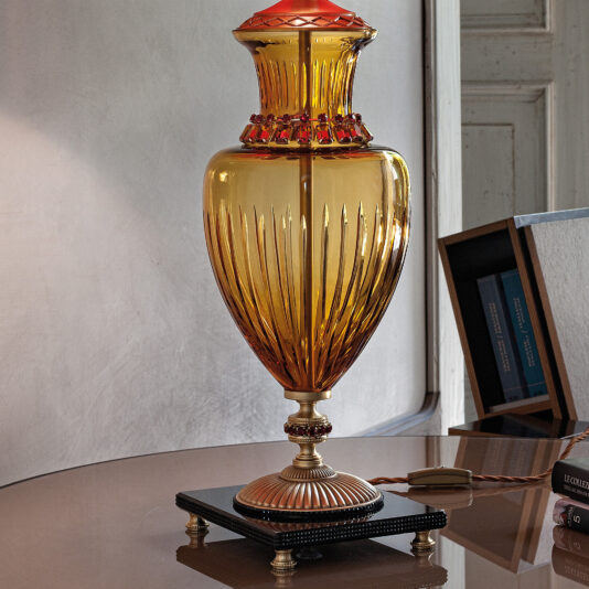 Classic Amber Glass Table Lamp With Ruby Stones A Classic Amber Glass Table Lamp With Ruby Stones, featuring an ornate amber glass body and a red-tinted top, sits on a decorative metal base atop a reflective surface. Behind it, there's a white wall and an open bookshelf filled with books. The lamp's elegant design enhances the scene's vintage aesthetic.